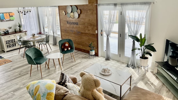 Una sala de estar moderna con un sofá, un sillón verde, una mesa de centro y un osito de peluche. Hay una pared de madera con un letrero de bienvenida y un comedor con una mesa y sillas.