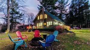 Una cabaña con terraza, rodeada de árboles y mobiliario exterior con sillas tipo Adirondack.