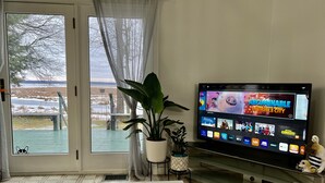 Una sala de estar con un televisor mostrando la interfaz de un servicio de streaming, una mesa de comedor con un control remoto y vistas a un paisaje nevado a través de una ventana.