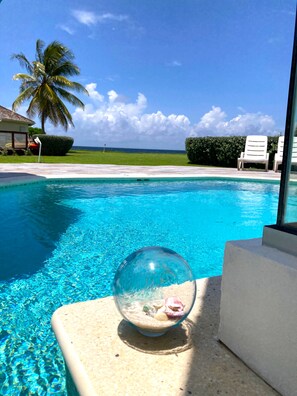 Une piscine d’un bleu éclatant, avec une sphère de verre contenant des coquillages, entourée de chaises longues et de palmiers.