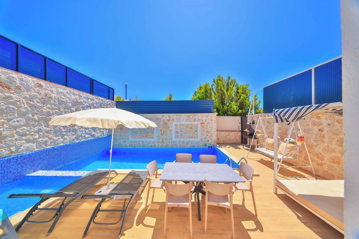 Área da piscina com mesa e cadeiras, uma espreguiçadeira e um pequeno espaço de estar com parede estampada.