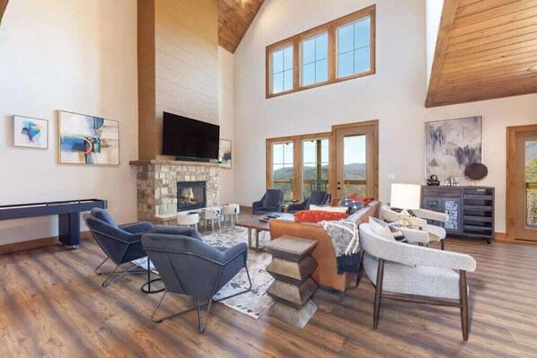 A spacious living room with a fireplace, multiple seating areas, and large windows offering views of the outdoors. - HUGE Mountain Retreat w/ POOL! + Arcades & View (Sevierville)