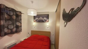 A bedroom with a red bedspread, a wall-mounted light fixture, a painting of mountains, and a wall decoration of a bird. - Charming T3 in a residence close to the slopes and shops South-facing balcony (La joue du loup)