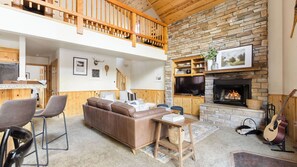 A spacious living area with a high ceiling, a stone fireplace, a television, and a guitar.