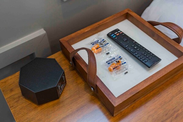 Una bandeja de madera con un control remoto, un altavoz y algunos artículos de tocador sobre una mesita de noche.