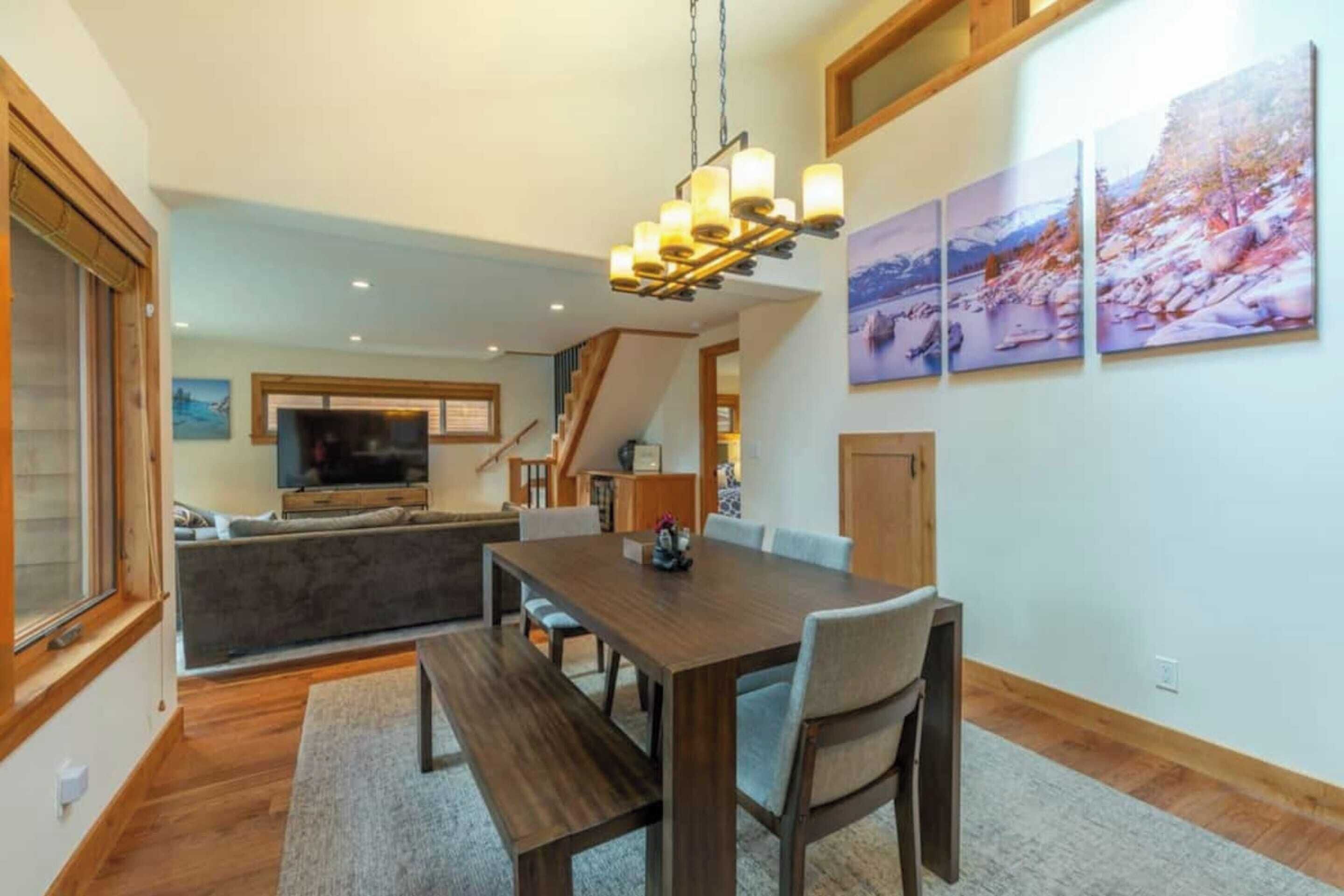 A dining area with a wooden table and benches, a chandelier, and a TV in the background.