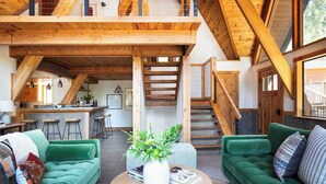 A spacious loft-style living area with a high ceiling and exposed wooden beams, a kitchen with a bar area, and a staircase leading to an upper level. - Timberline by Avantstay Stunning Chalet Cabin w/ Hot Tub, Pool Table & Home Theater! (Carnelian Bay)