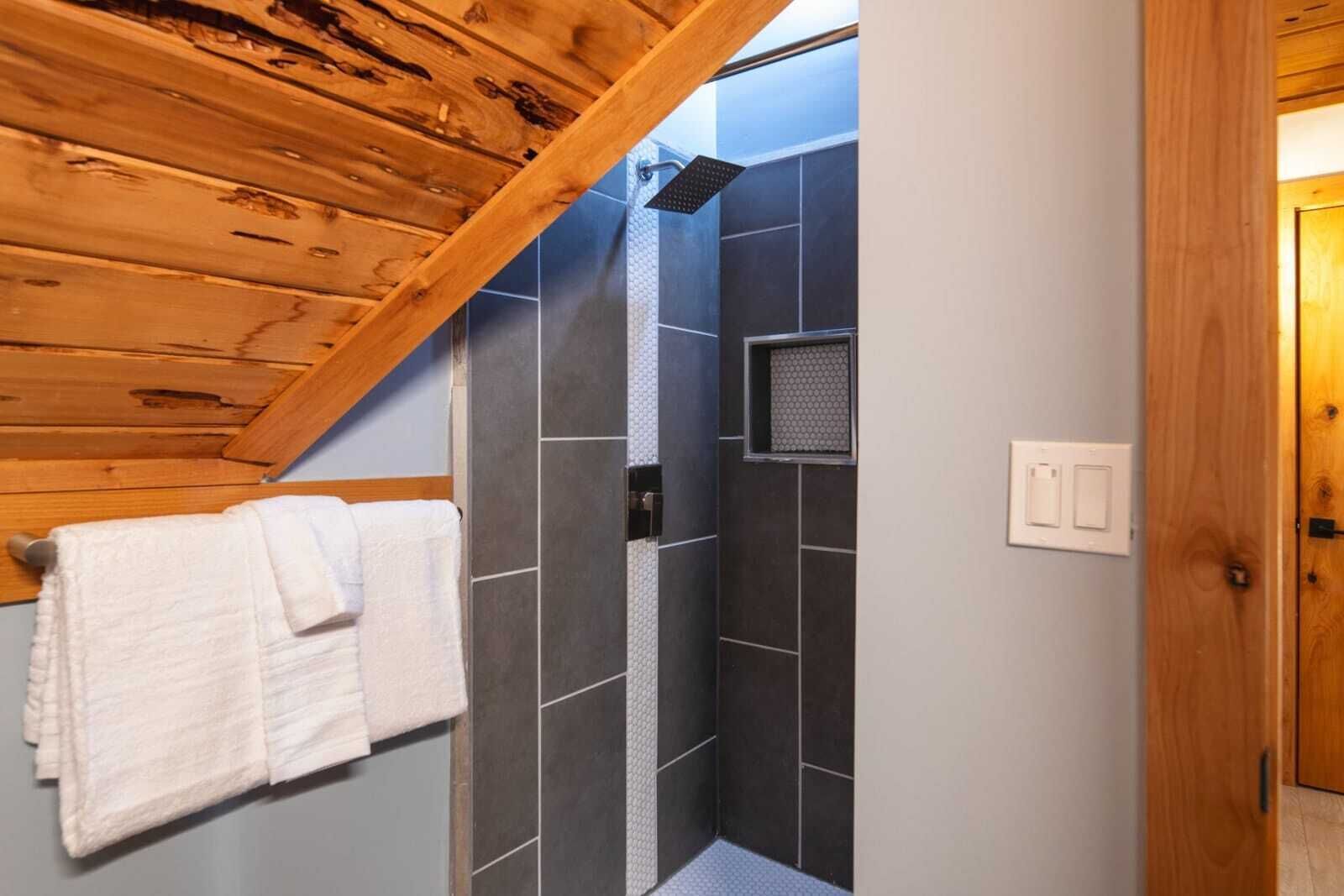 A shower area with dark tiles, a wooden ceiling, and a towel rack with white towels.