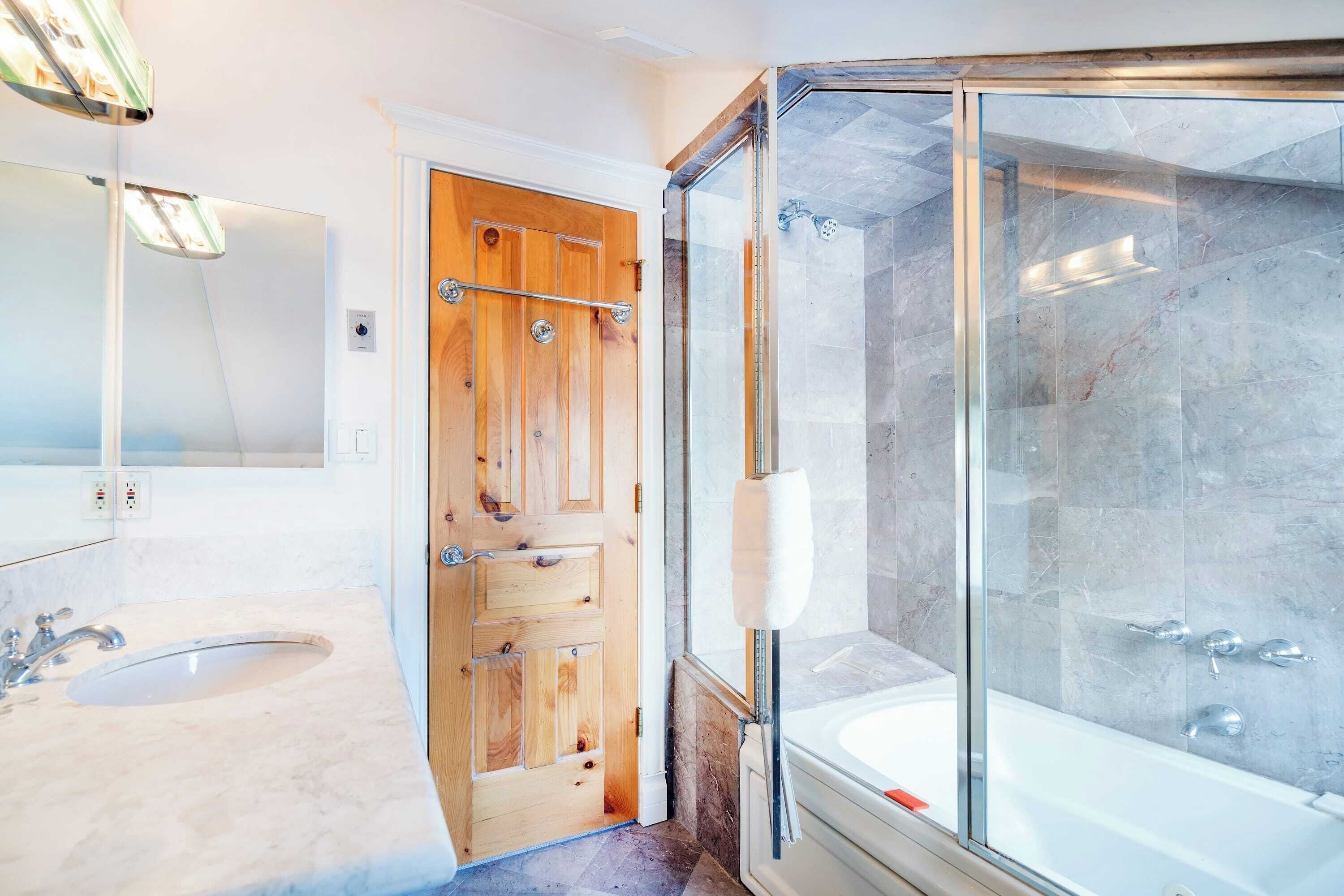 A modern bathroom with a wooden door, a glass shower enclosure, and a marble sink.