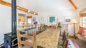 Een knus vertrek met een houten eettafel, stoelen, een zwart fornuis, houten balken, een vensterbank en een keukenhoek.