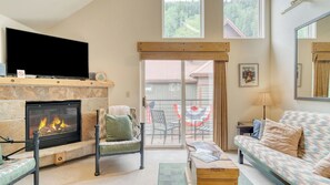 Una sala de estar con chimenea, un sofá, un sillón, un televisor y vistas a un balcón con mobiliario exterior.