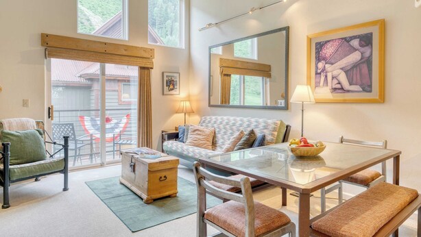 A cozy living room with a sofa, a dining table, and chairs. There is a window with curtains, a lamp, and a painting on the wall.