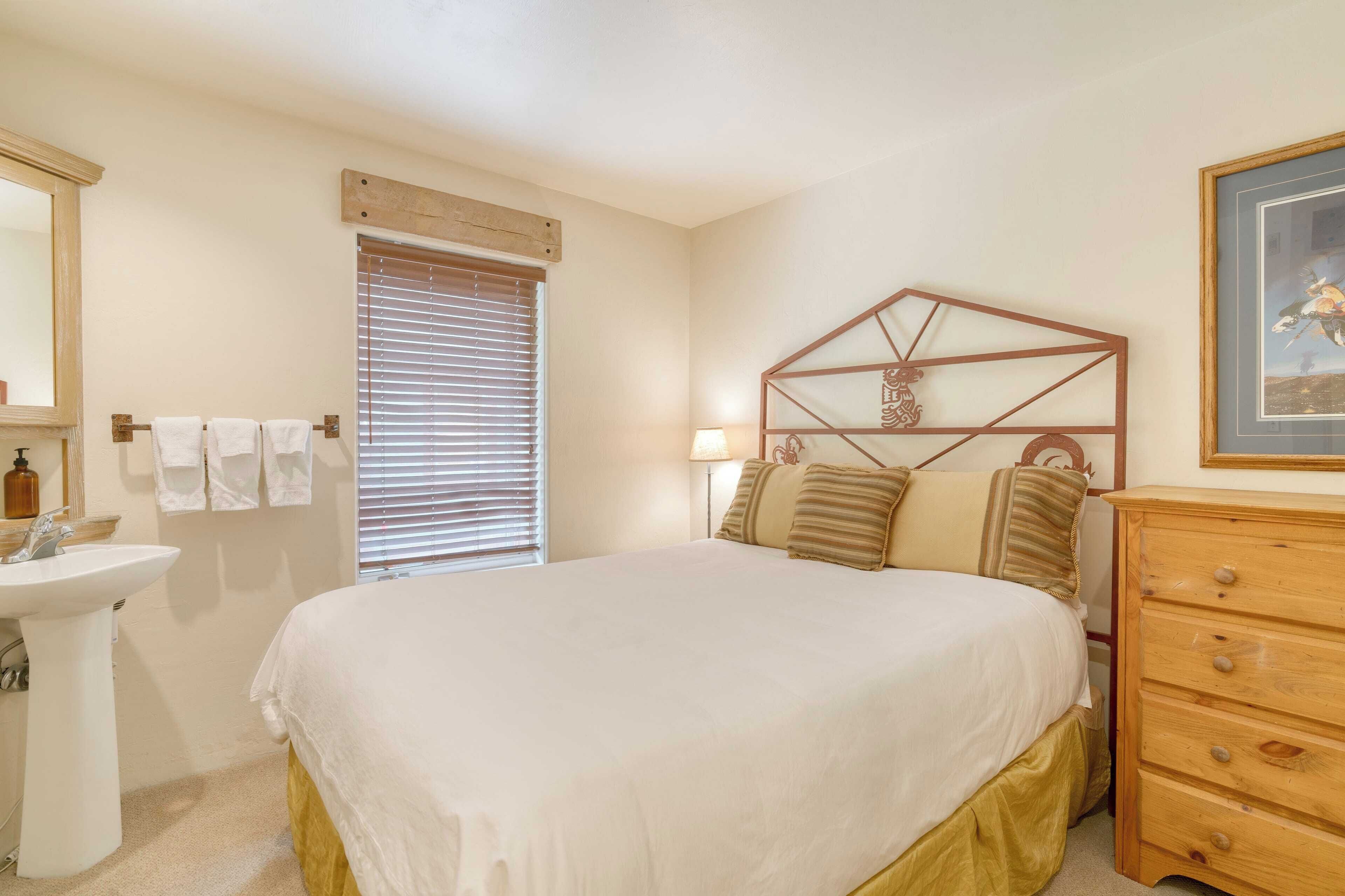 A bedroom with a bed, a wooden dresser, a bedside lamp, a window with blinds, and a bathroom with a sink and towel rack.