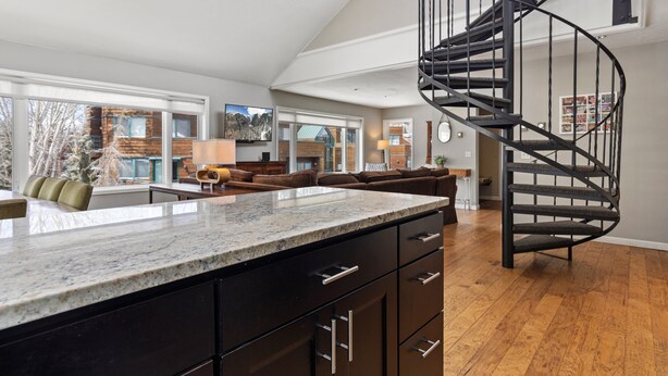 A spacious living area with a kitchen island, a spiral staircase, and a large window overlooking a brick building. - Ghostriders 10 by Avantstay Penthouse Suite w/ Large Patio & Mountain Views! (Telluride)