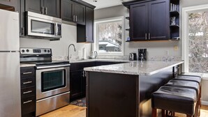 A modern kitchen with dark cabinets, stainless steel appliances, a large island with bar stools, and a window providing a view of the outdoors. - Ghostriders 10 by Avantstay Penthouse Suite w/ Large Patio & Mountain Views! (Telluride)
