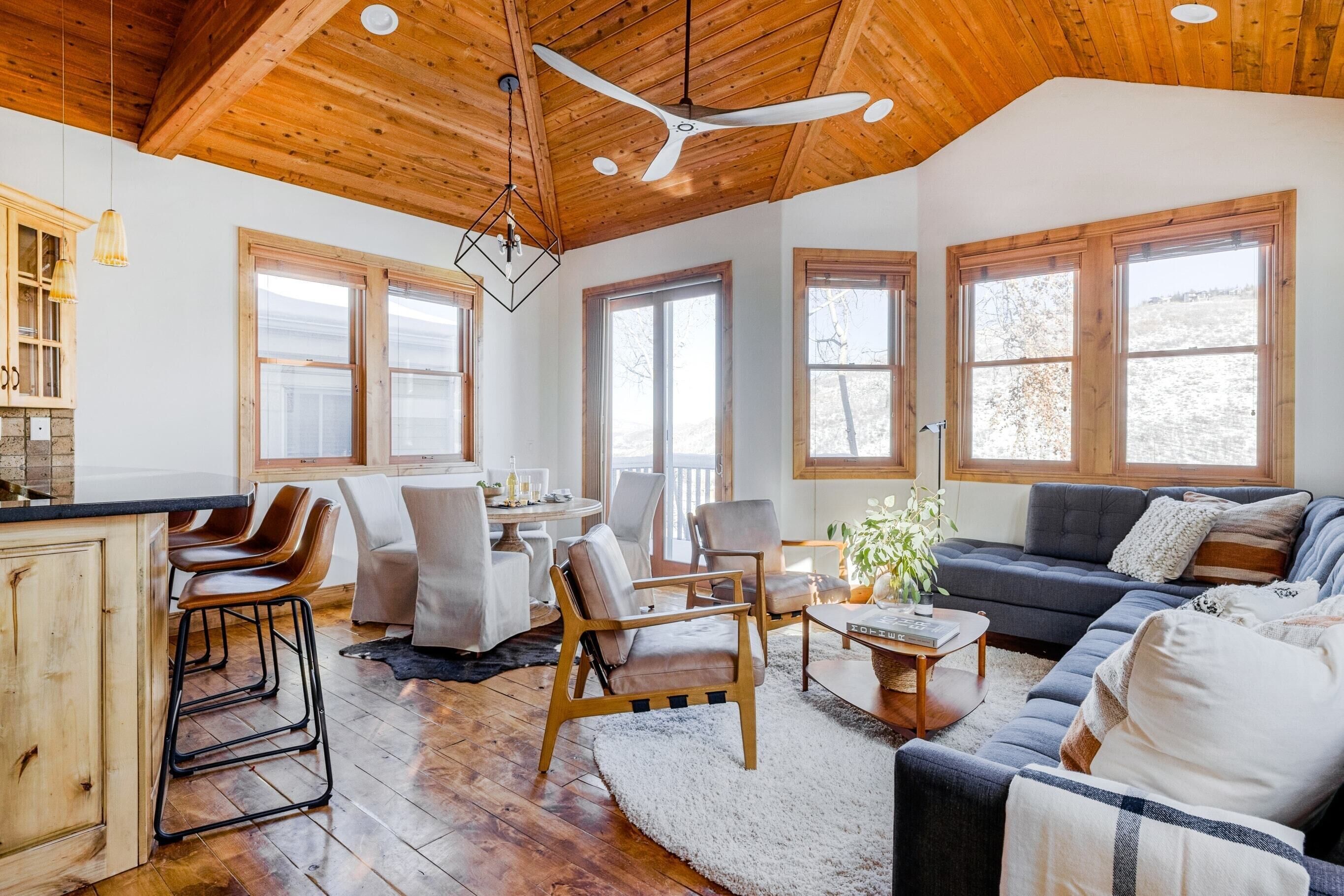 A spacious living area with a wooden ceiling, large windows, a sofa, chairs, and a kitchen area with bar stools.
