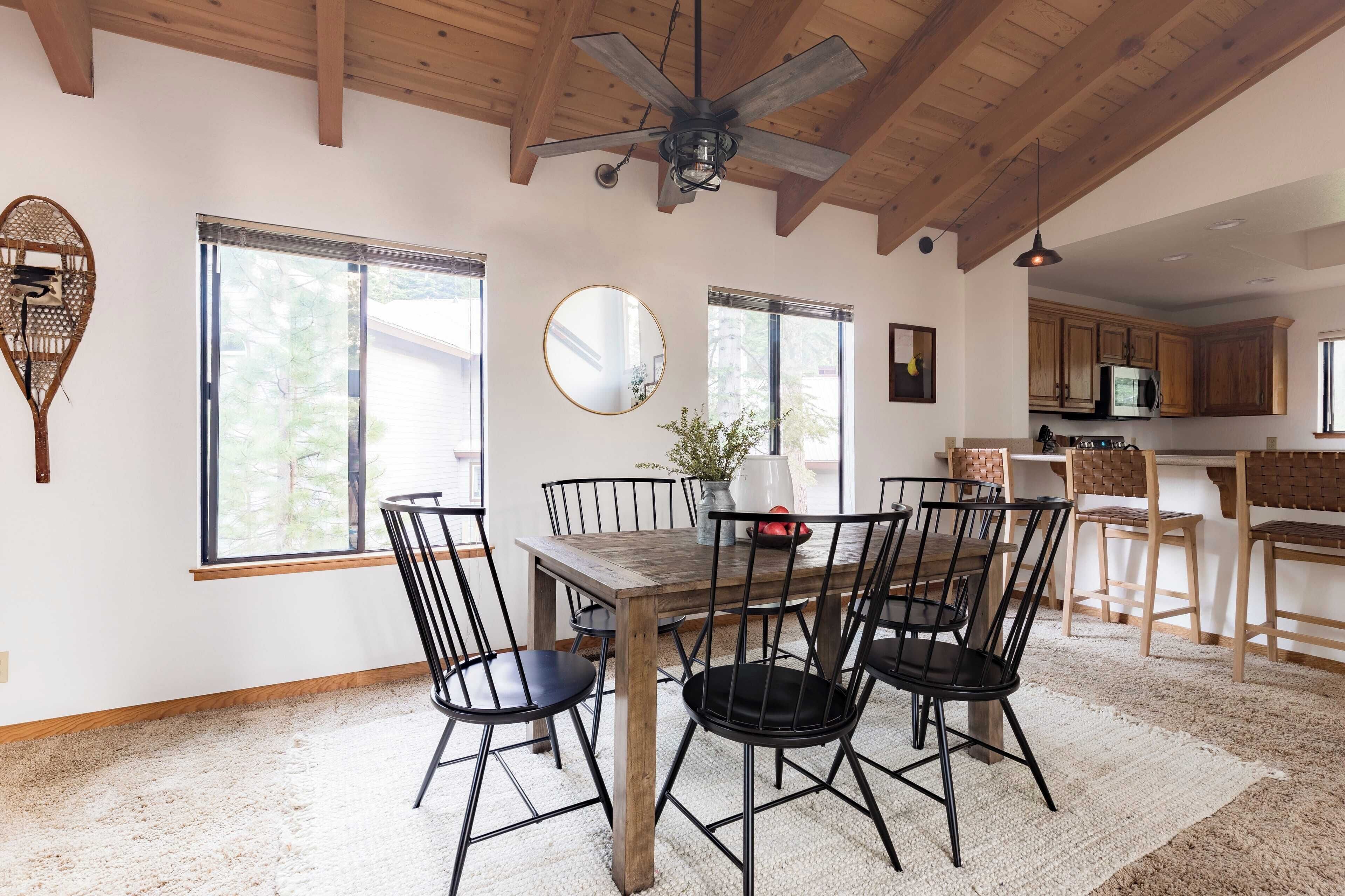 Un coin repas avec une table et des chaises en bois, un ventilateur de plafond, de grandes fenêtres et une cuisine en arrière-plan.
