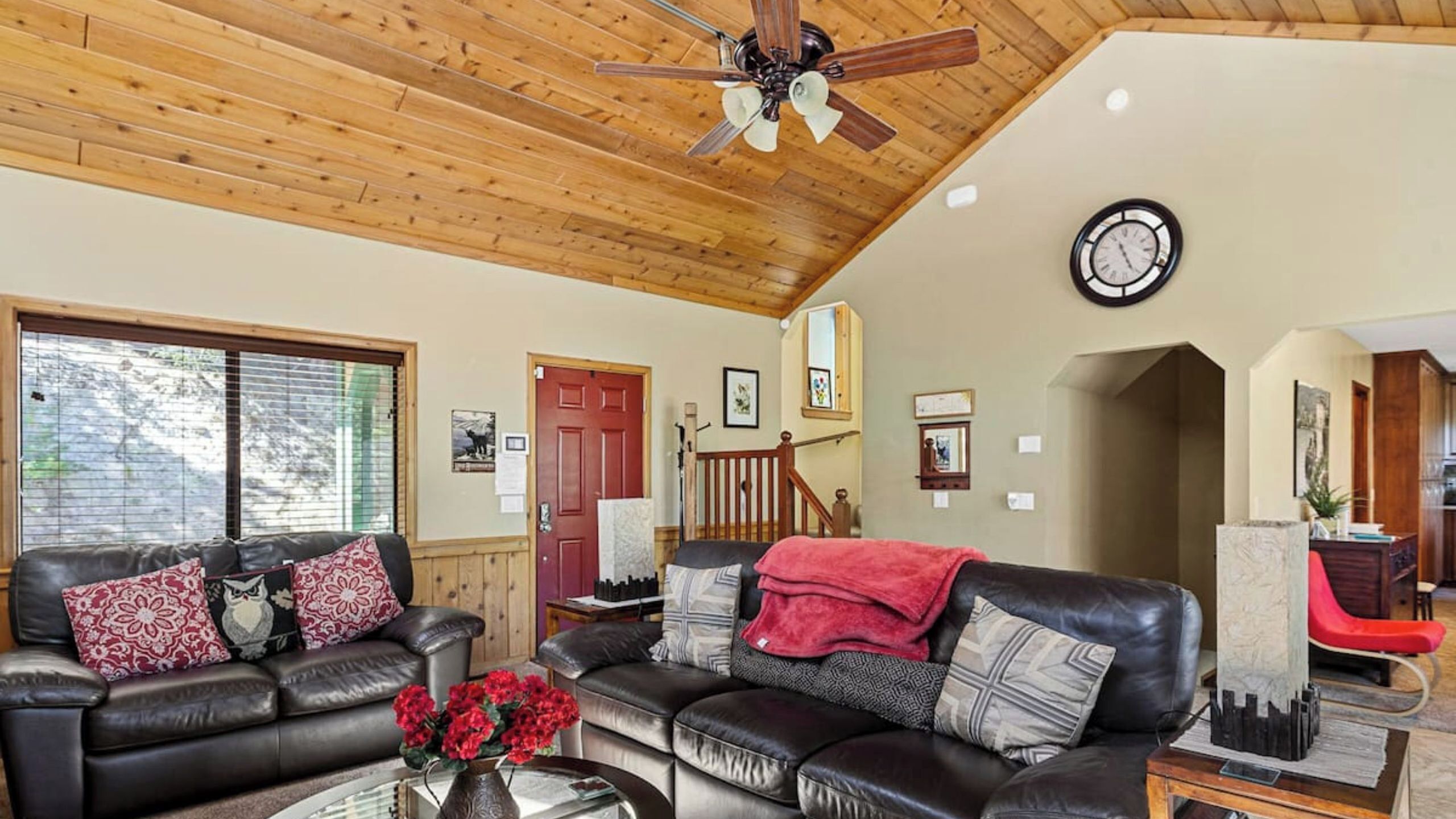 A spacious living room with a wooden ceiling, a black leather sofa, a red throw blanket, and a clock on the wall.