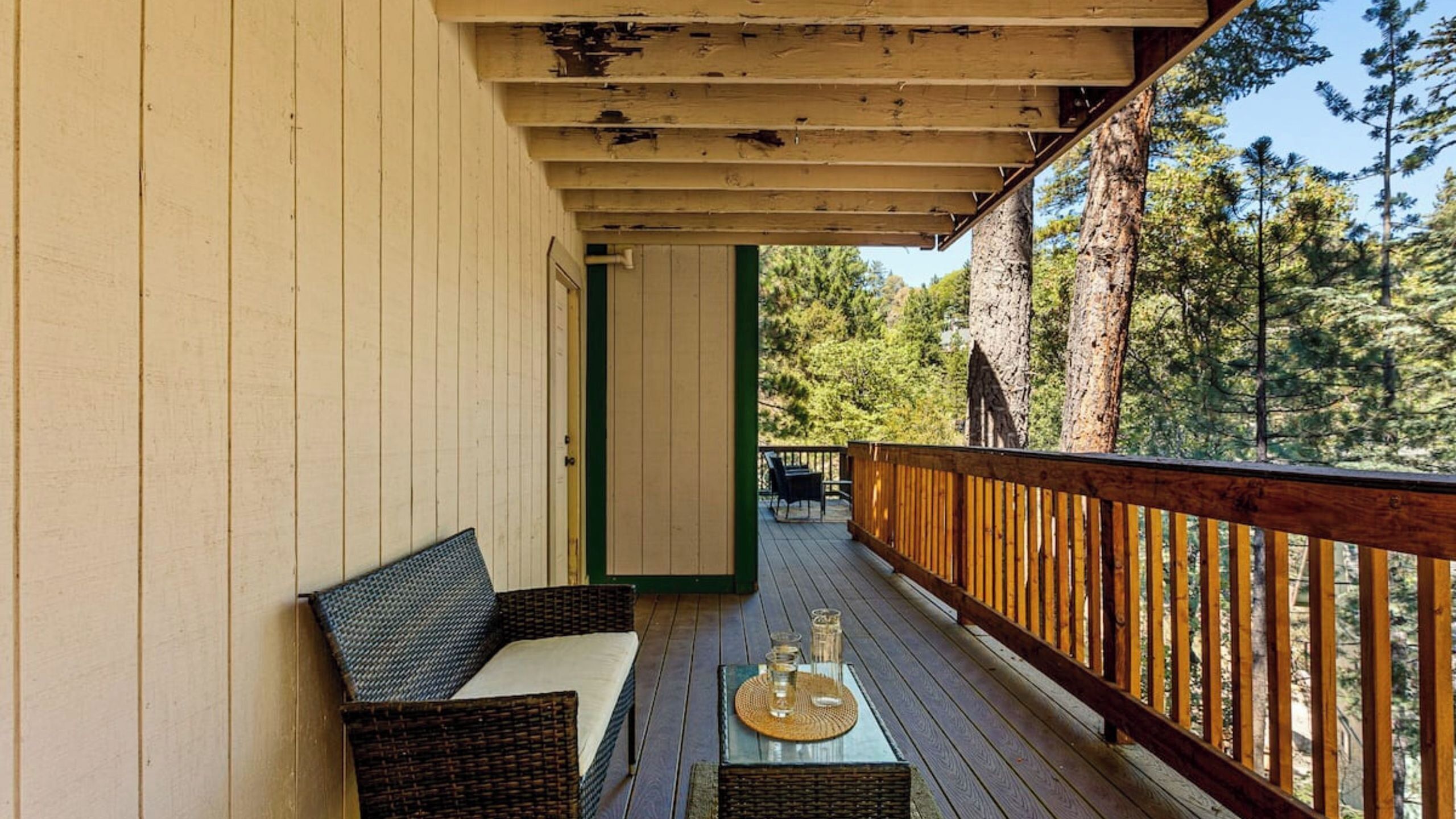 A wooden deck with a sofa and a table, surrounded by a wooden railing, overlooking a forest.