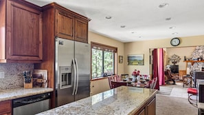 A kitchen with wooden cabinets, a stainless steel refrigerator, granite countertops, a dining area with a table and chairs, and a stone fireplace. - Beautifully Remodeled Kitchen Peaceful Mountain Views Sutter by Avantstay (Lake Arrowhead)