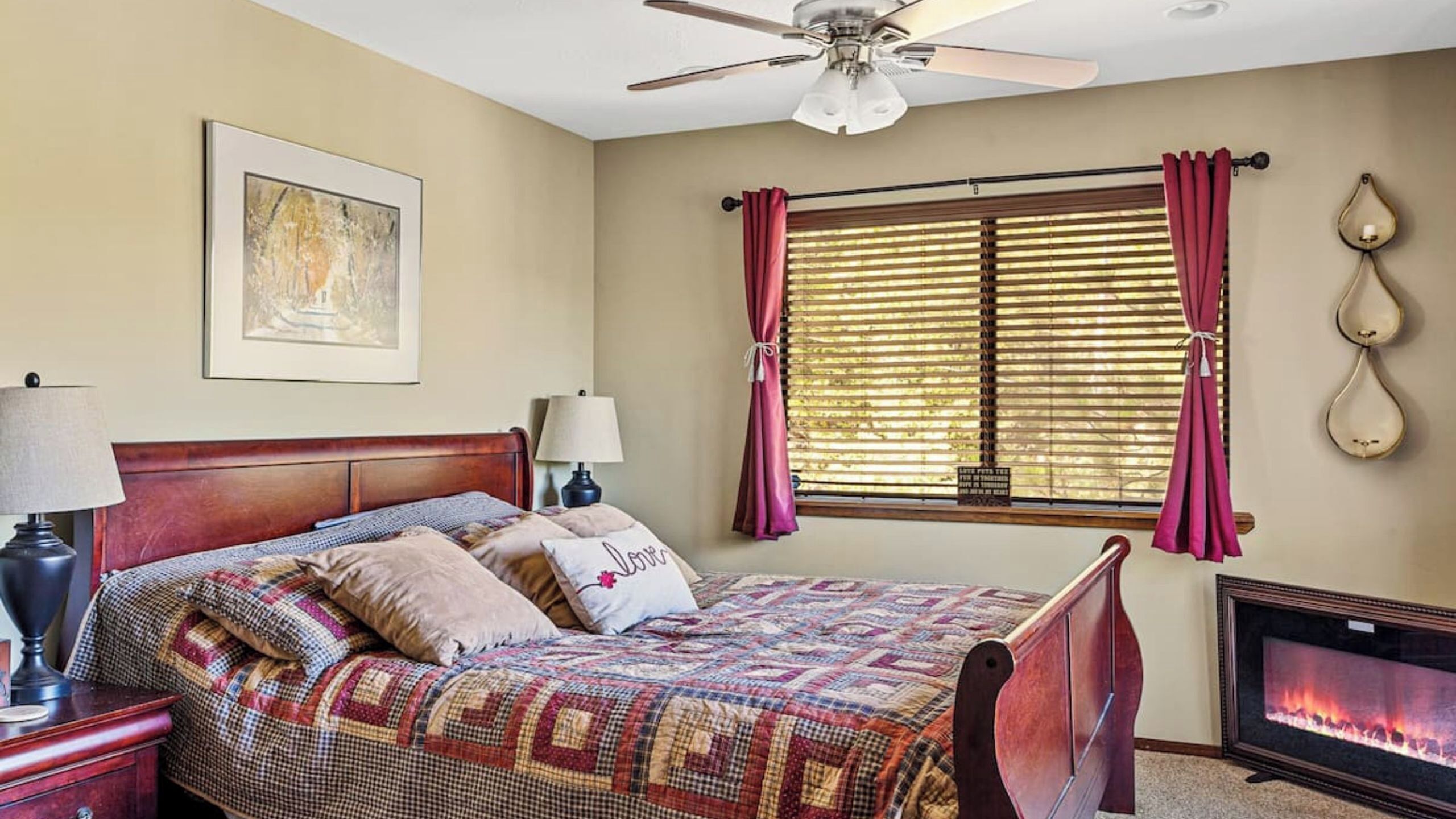 Un dormitorio con cama, ventilador de techo, chimenea, ventana con cortinas e imagen enmarcada en la pared.