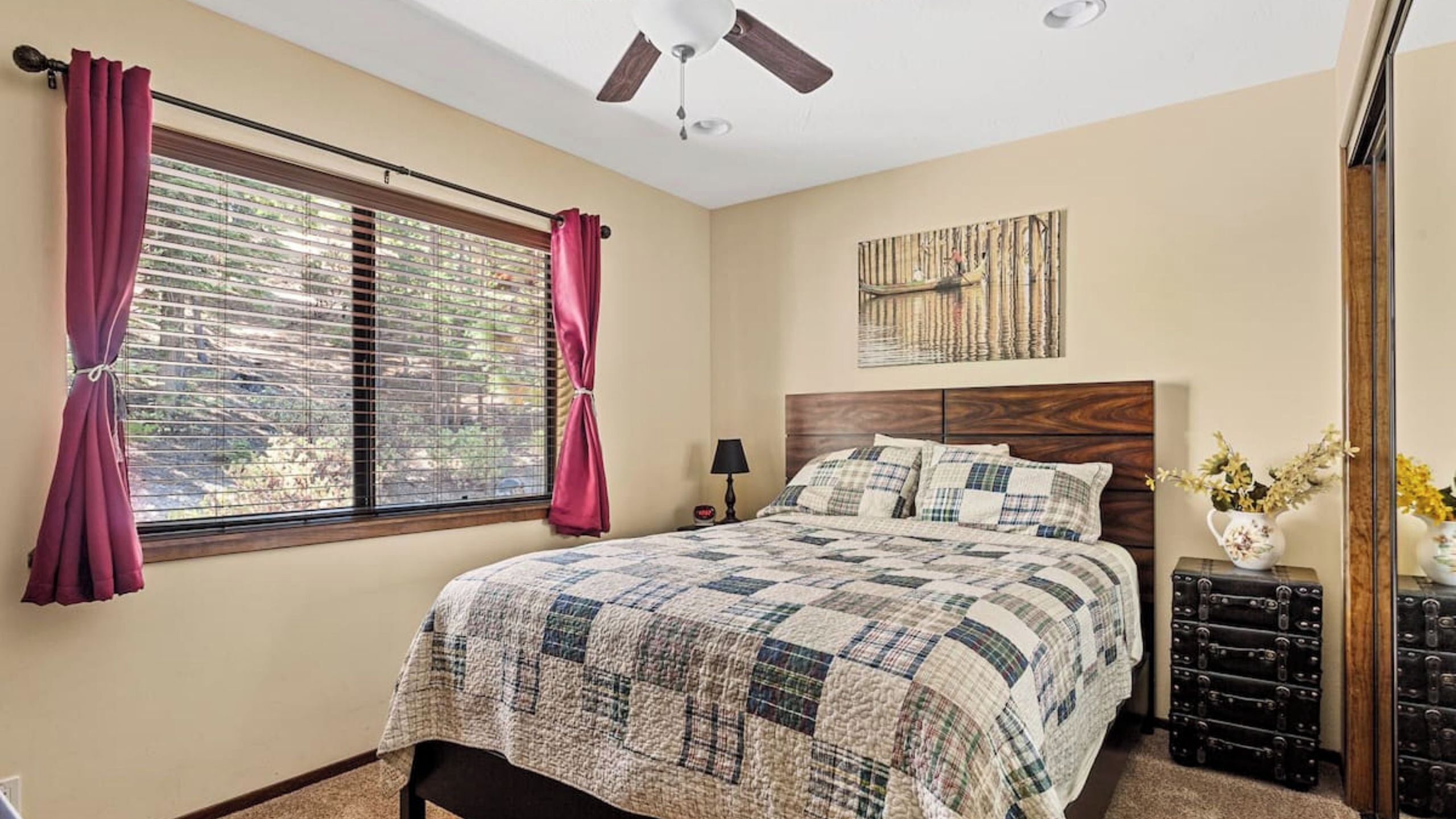 A bedroom with a bed, a ceiling fan, a window with blinds, a nightstand, a lamp, and a wall-mounted artwork.