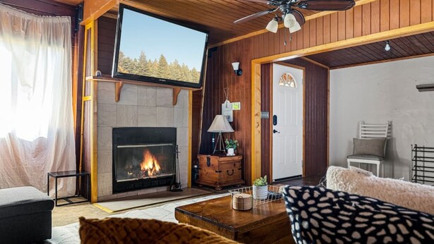 Ein gemütliches Wohnzimmer mit Kamin, Holzwänden, Deckenventilator und großem Fenster mit Blick auf einen Wald.