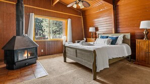 Ein Schlafzimmer einer Holzhütte mit Bett, Nachttisch, Lampe, Fenster mit Vorhängen, Kamin und Deckenventilator.
