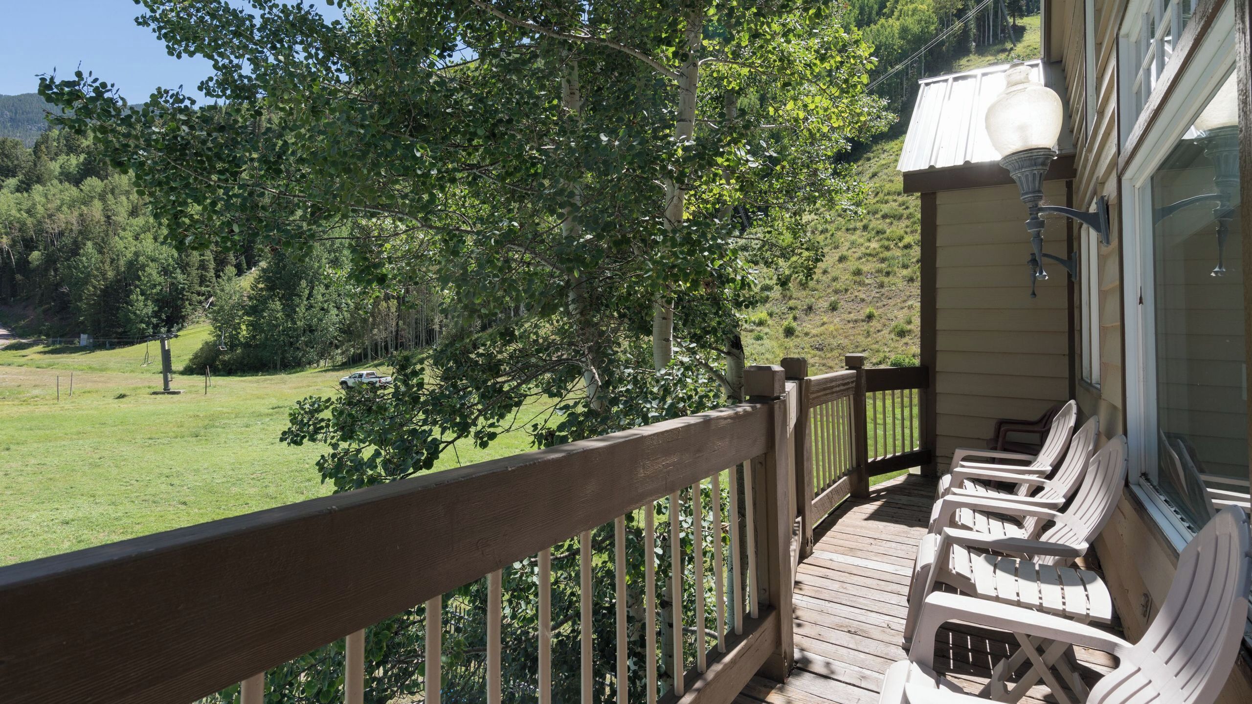 Terraza de madera con mobiliario exterior, con vistas a una zona verde con árboles y una colina distante.