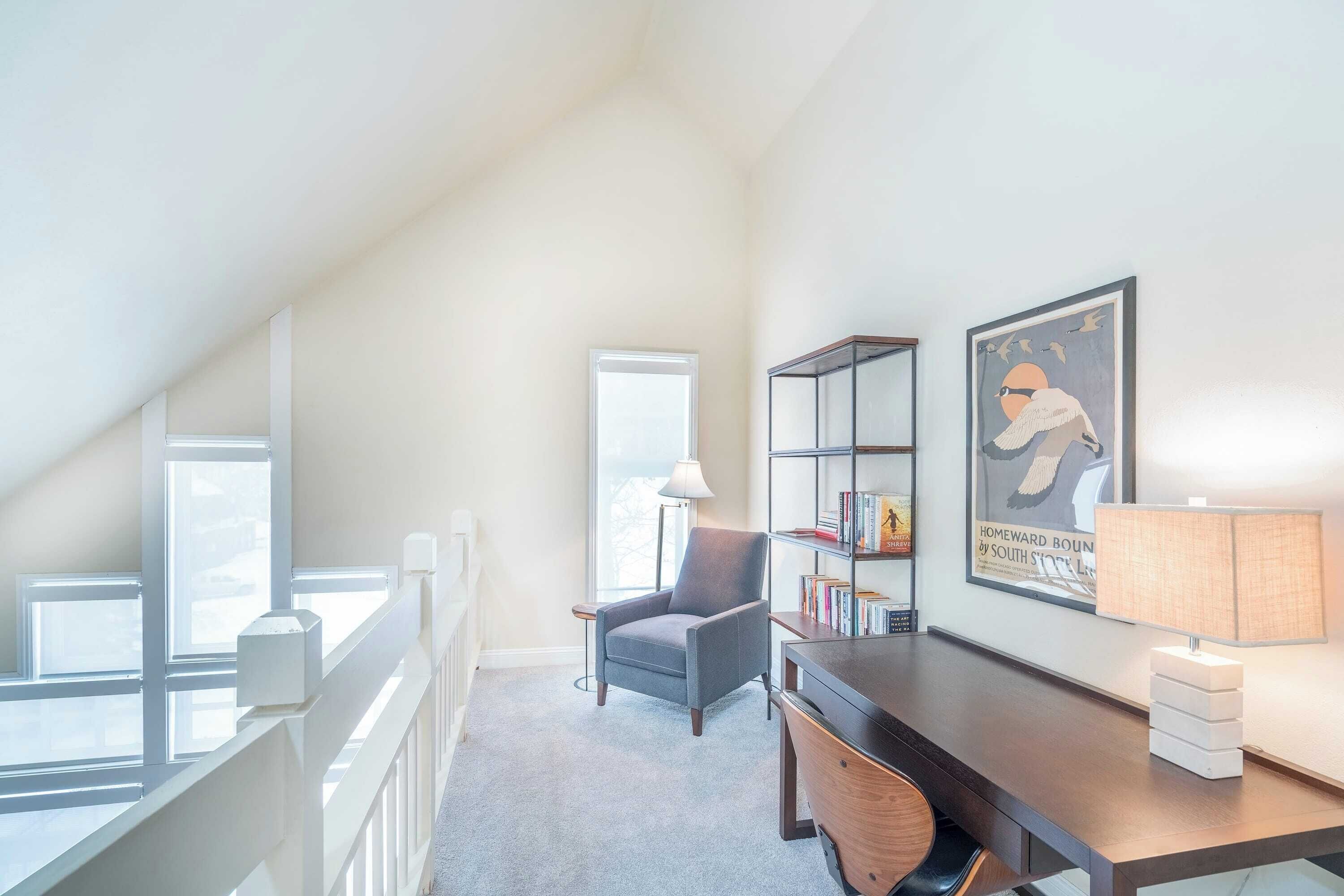 A high-ceilinged room with a desk, chair, bookshelf, and a framed picture on the wall.