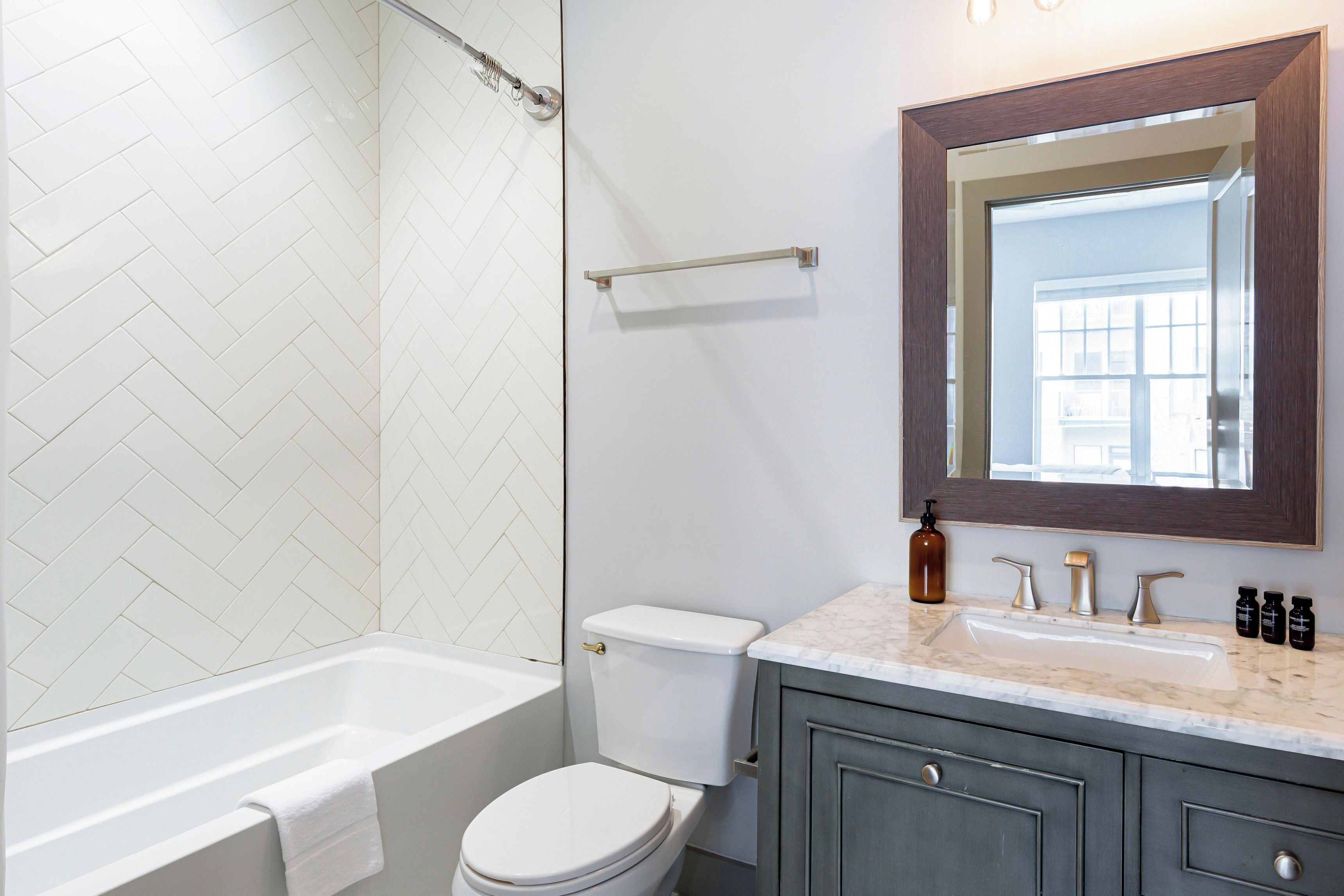 Um banheiro com parede de azulejos brancos em padrão herringbone, vaso sanitário branco, banheira, bancada de mármore, espelho e porta-toalhas.