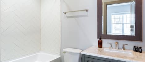 Um banheiro com parede de azulejos brancos em padrão herringbone, vaso sanitário branco, banheira, bancada de mármore, espelho e porta-toalhas.