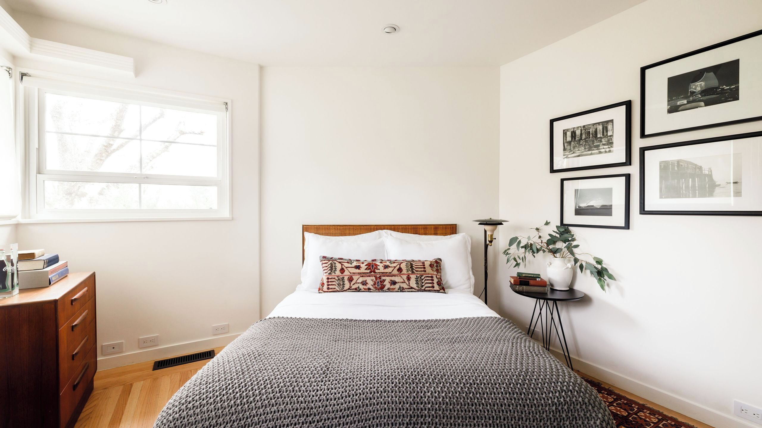 A bedroom with a bed, a bedside table, a lamp, a window, and framed pictures on the wall.