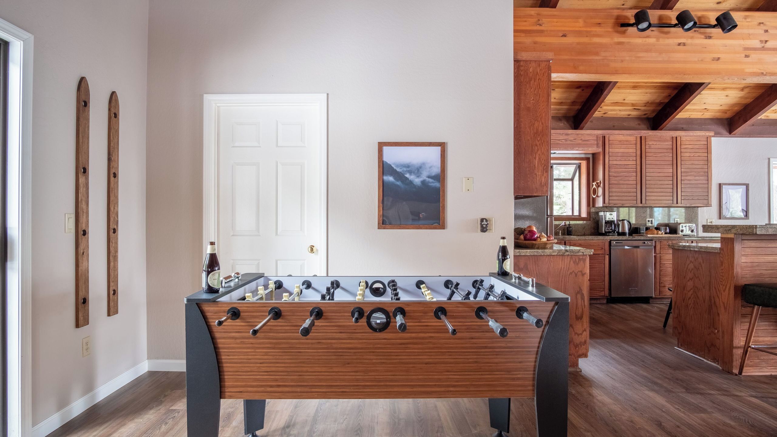 A room with a foosball table, wooden flooring, a kitchen with wooden cabinets, and a white door.