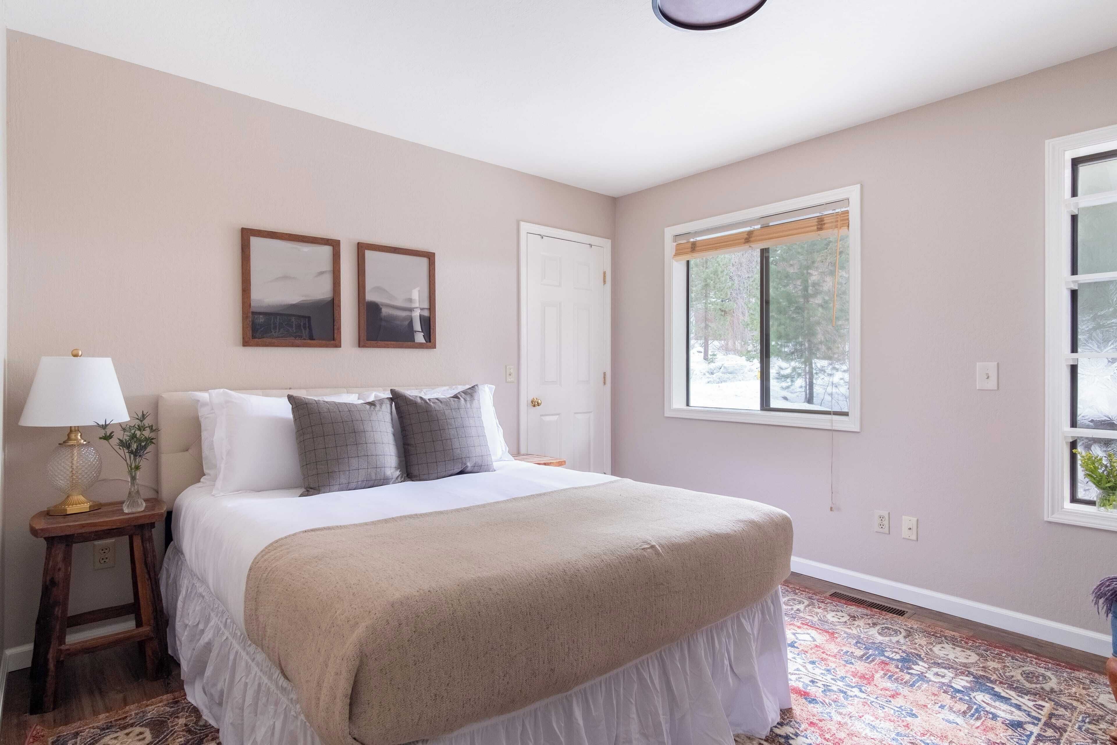 A bedroom with a bed, a nightstand, a lamp, two framed pictures, a door, and a window with a view of trees.