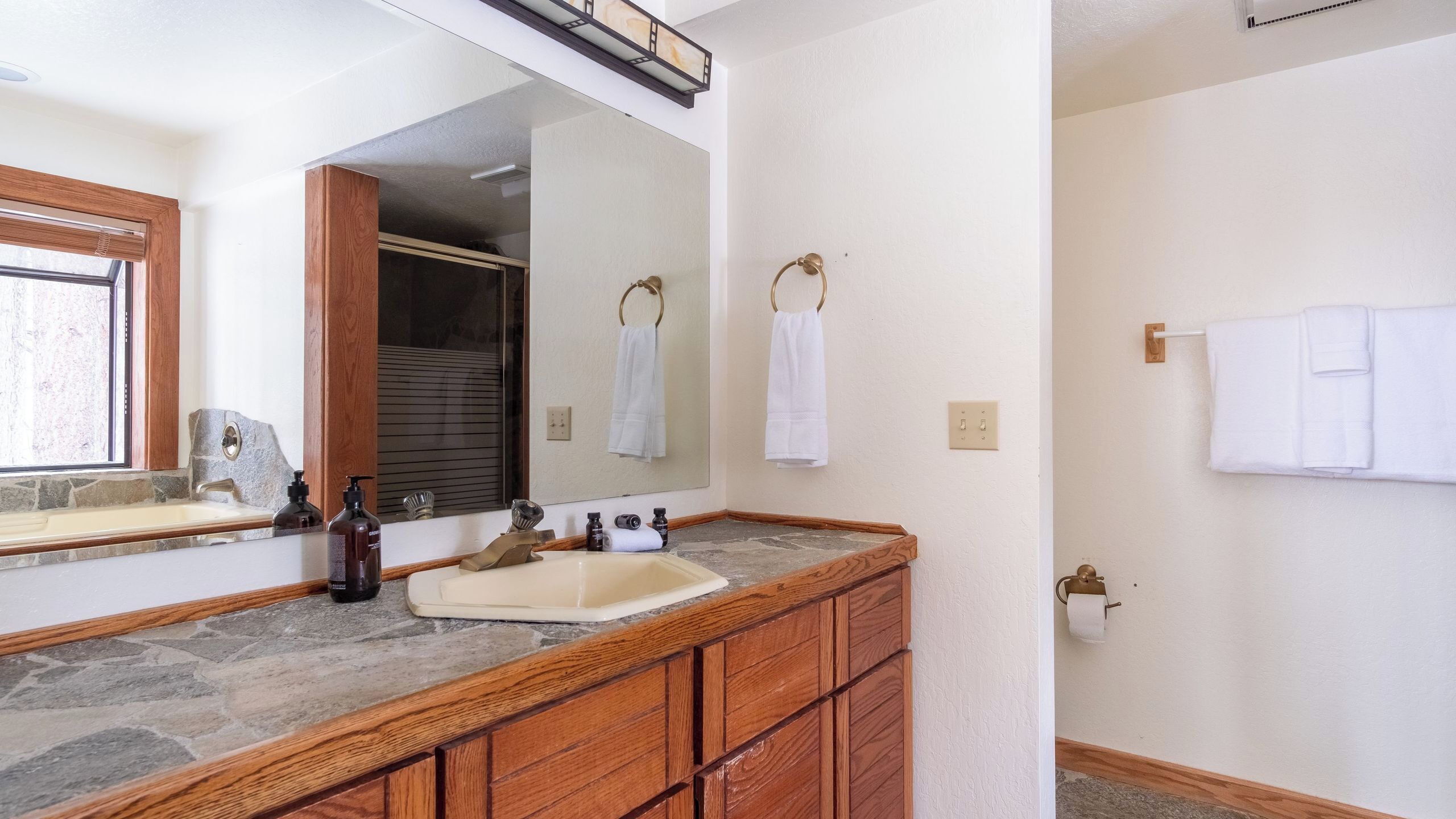 A bathroom with a stone countertop, wooden cabinets, a large mirror, and two white towels hanging on hooks.
