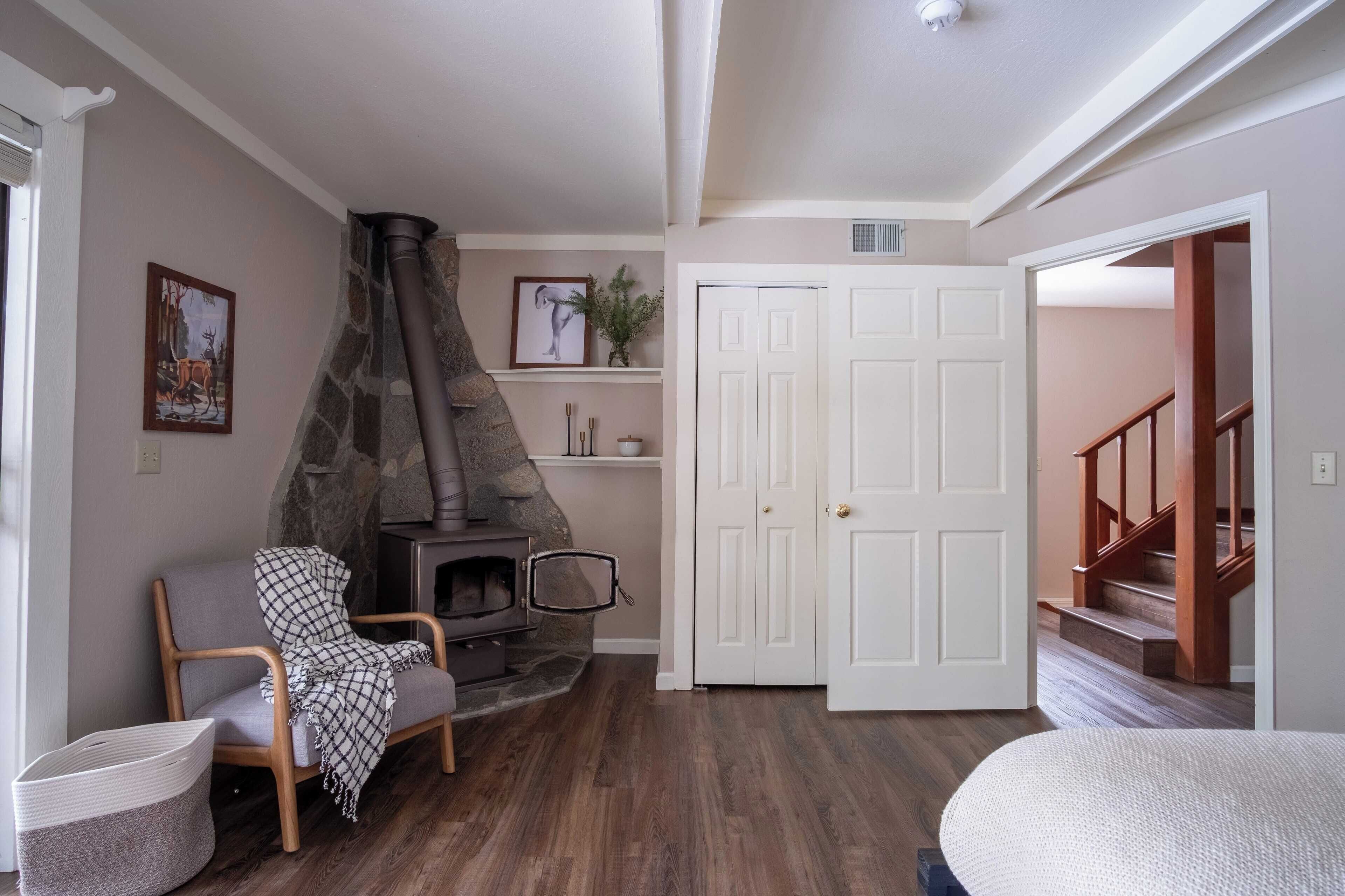 A cozy living room with a stone fireplace, a comfortable chair, a white door, and a staircase leading to another level.