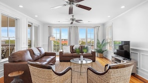 A spacious living room with a brown leather sofa, a glass coffee table, wicker chairs, a ceiling fan, and a television.
