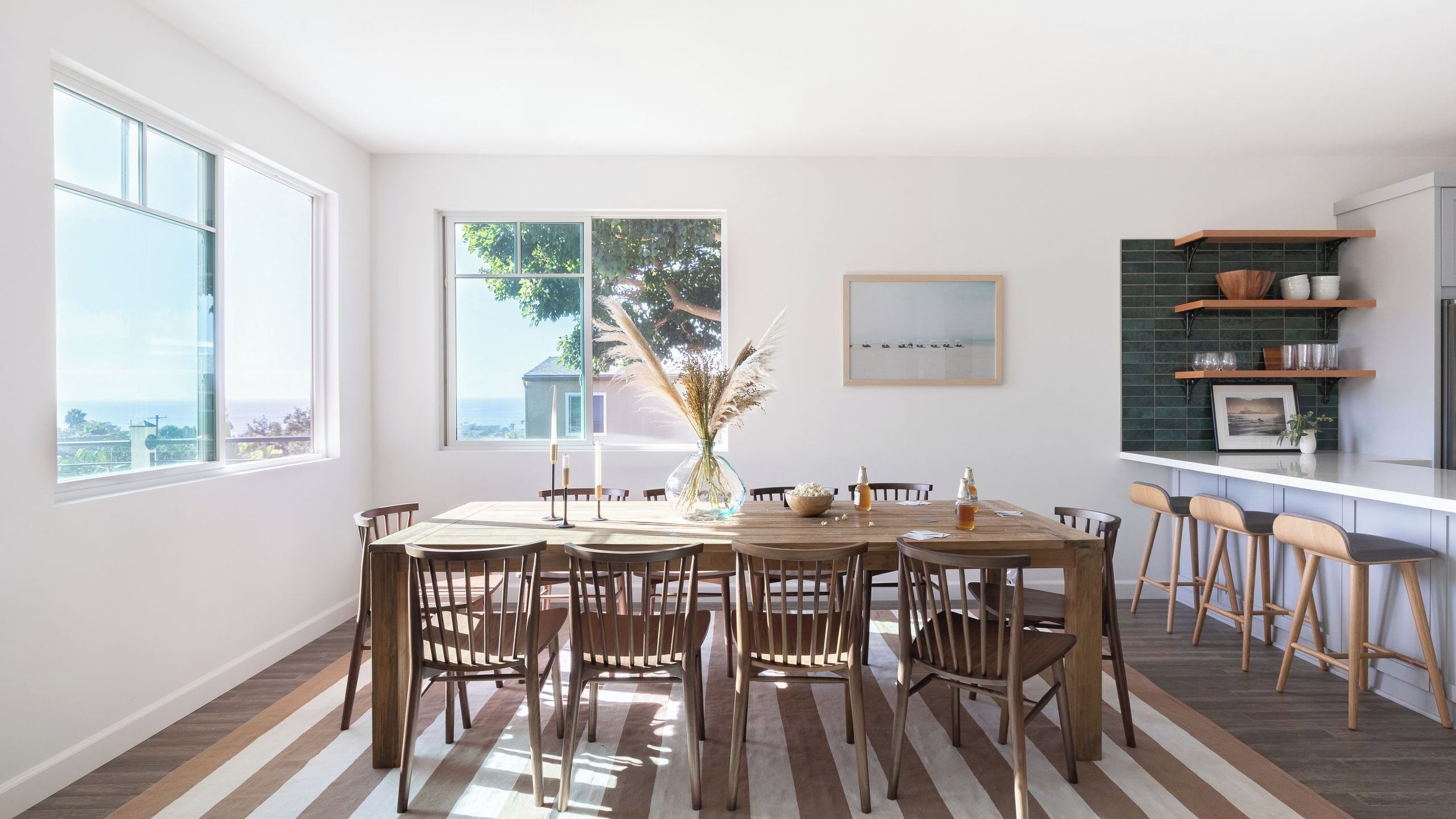 A dining area with a wooden table and chairs, a vase with pampas grass, and a view of the ocean through large windows.