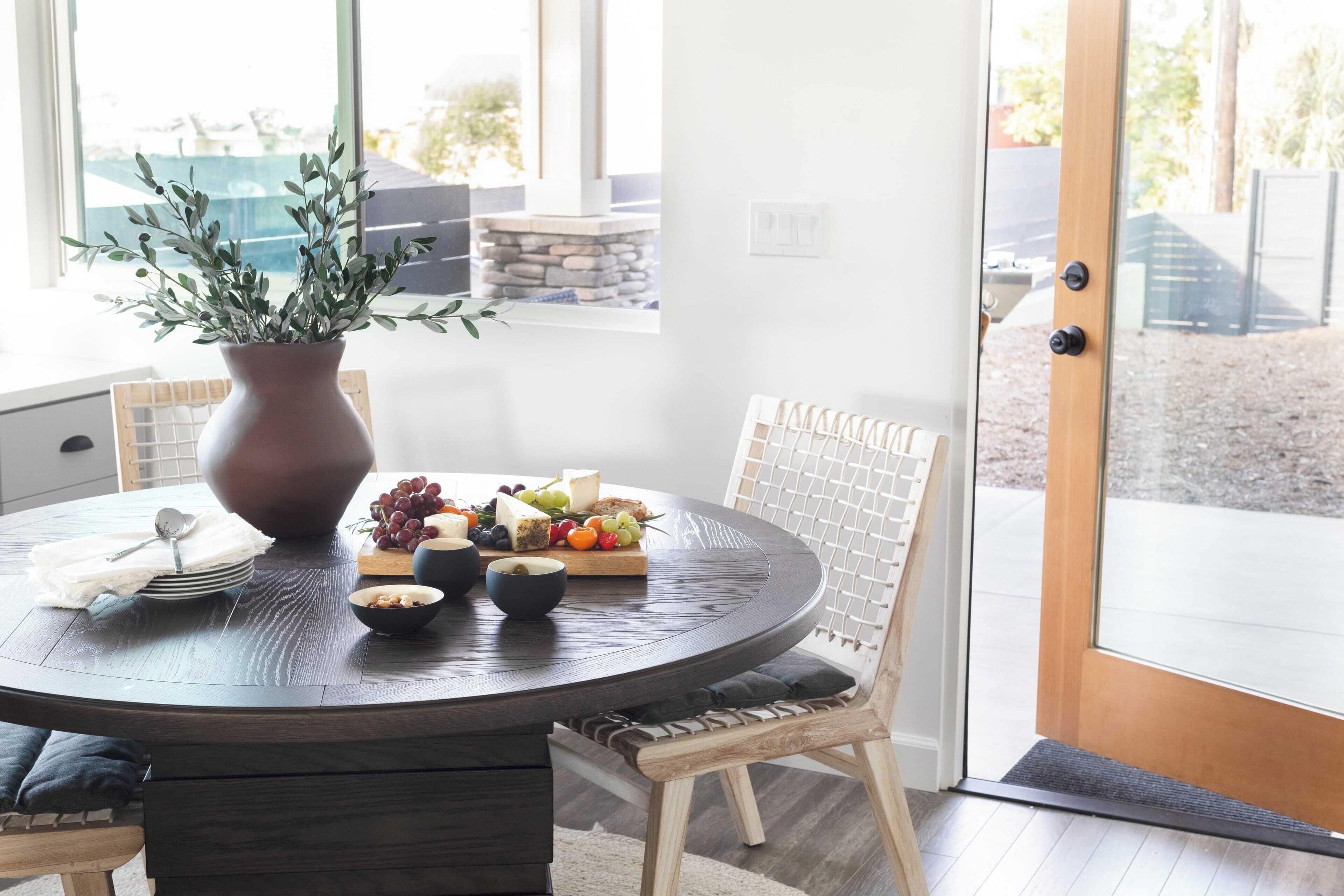 A dining area with a round table, a vase with greenery, and a fruit and cheese board.