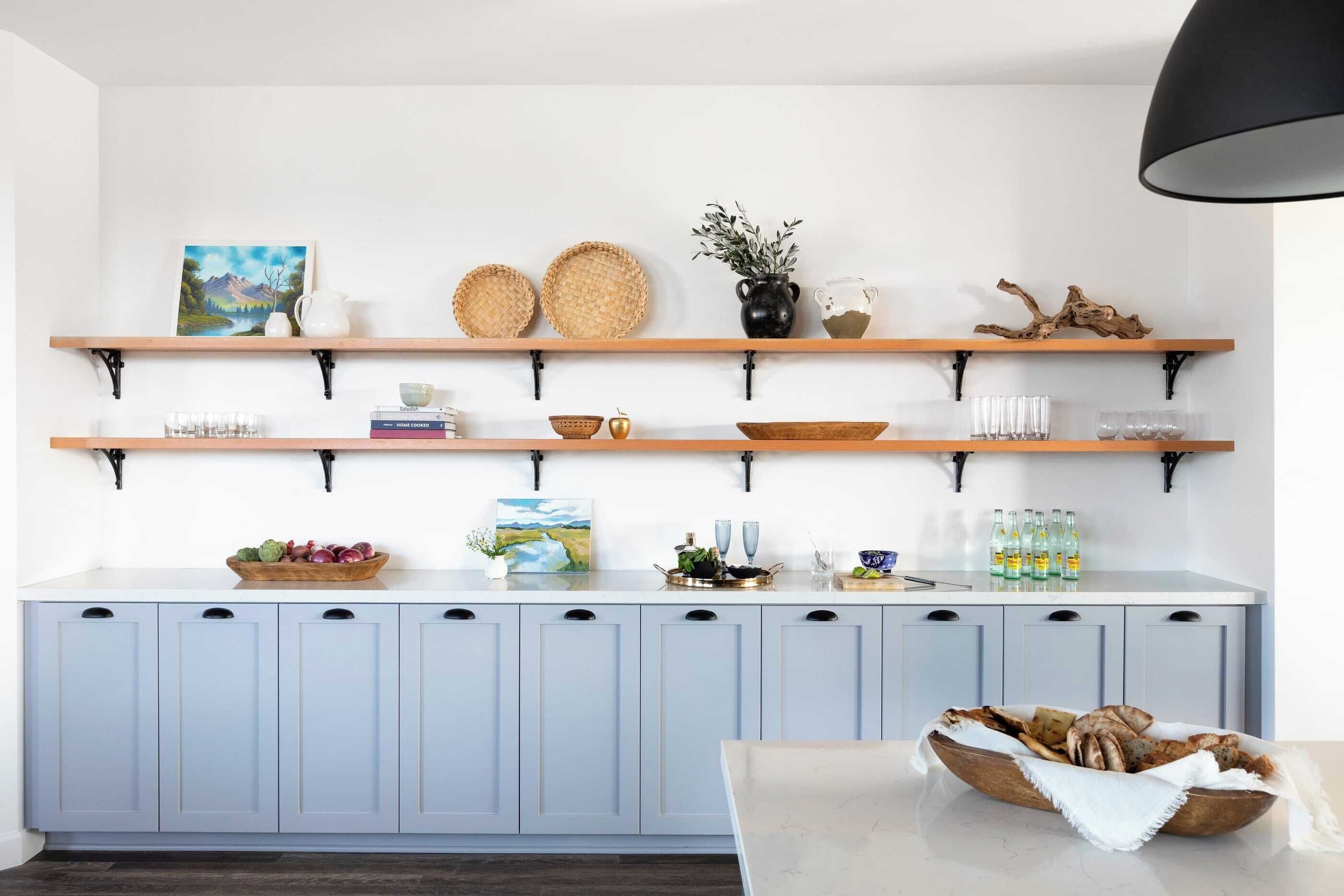 Una cocina con armarios azul claro, repisas de madera con diversos utensilios de cocina y una mesa de comedor con pan.