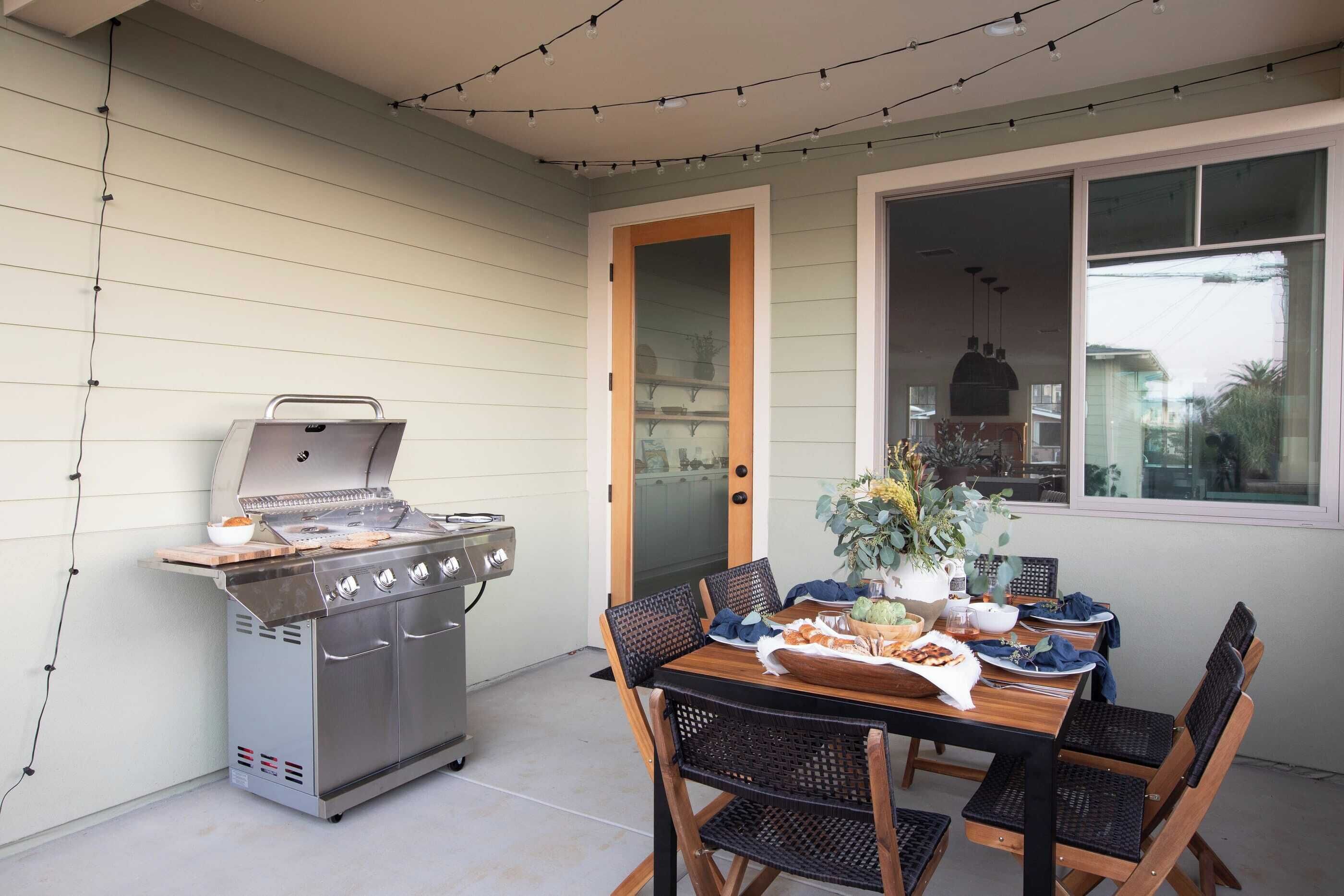 Un espace extérieur aménagé pour les repas, avec un barbecue, une table, des chaises et des guirlandes lumineuses.