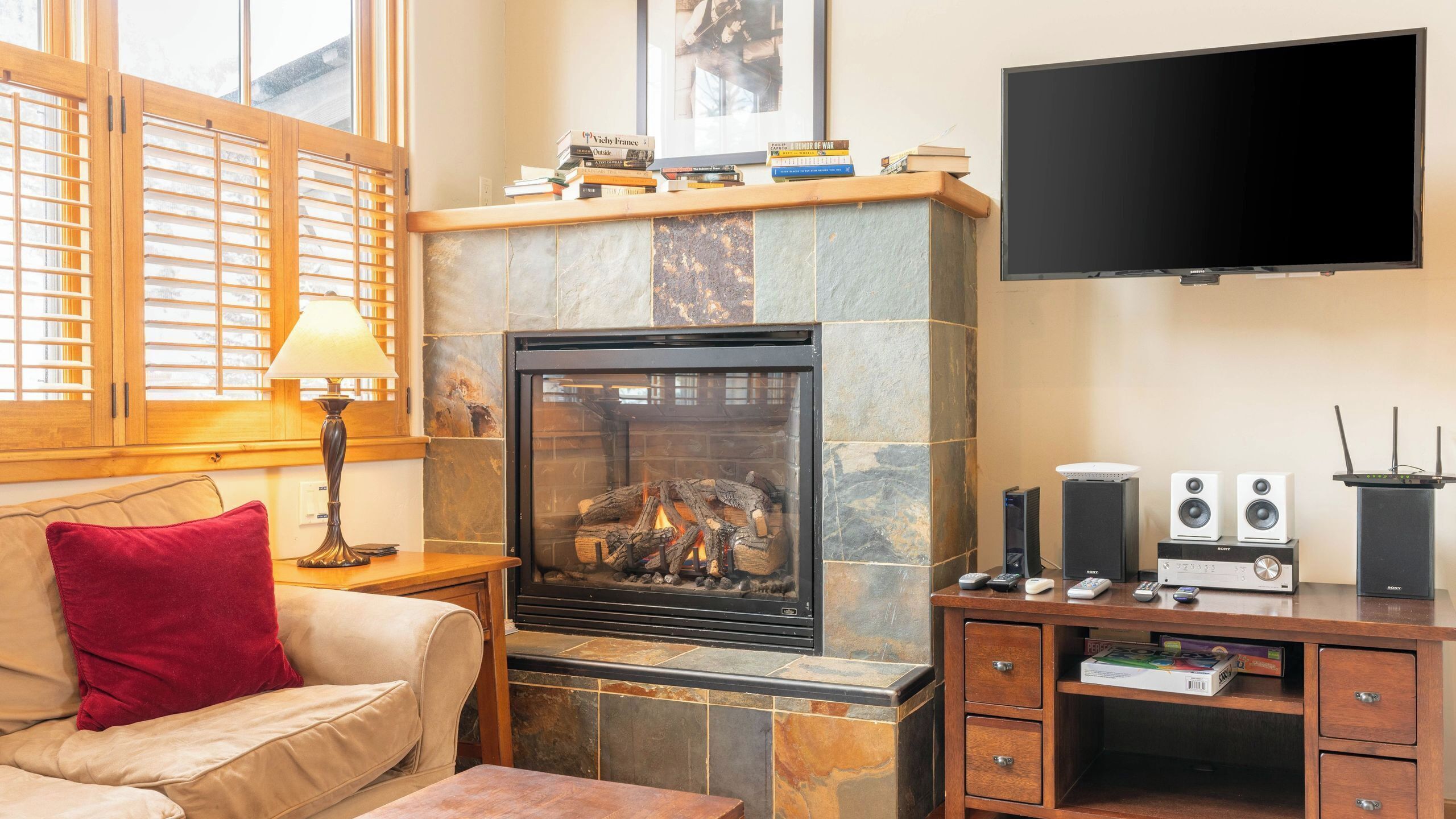 A cozy living room with a fireplace, a sofa with a red pillow, a TV mounted on the wall, and a wooden entertainment unit with speakers and a Sony TV.