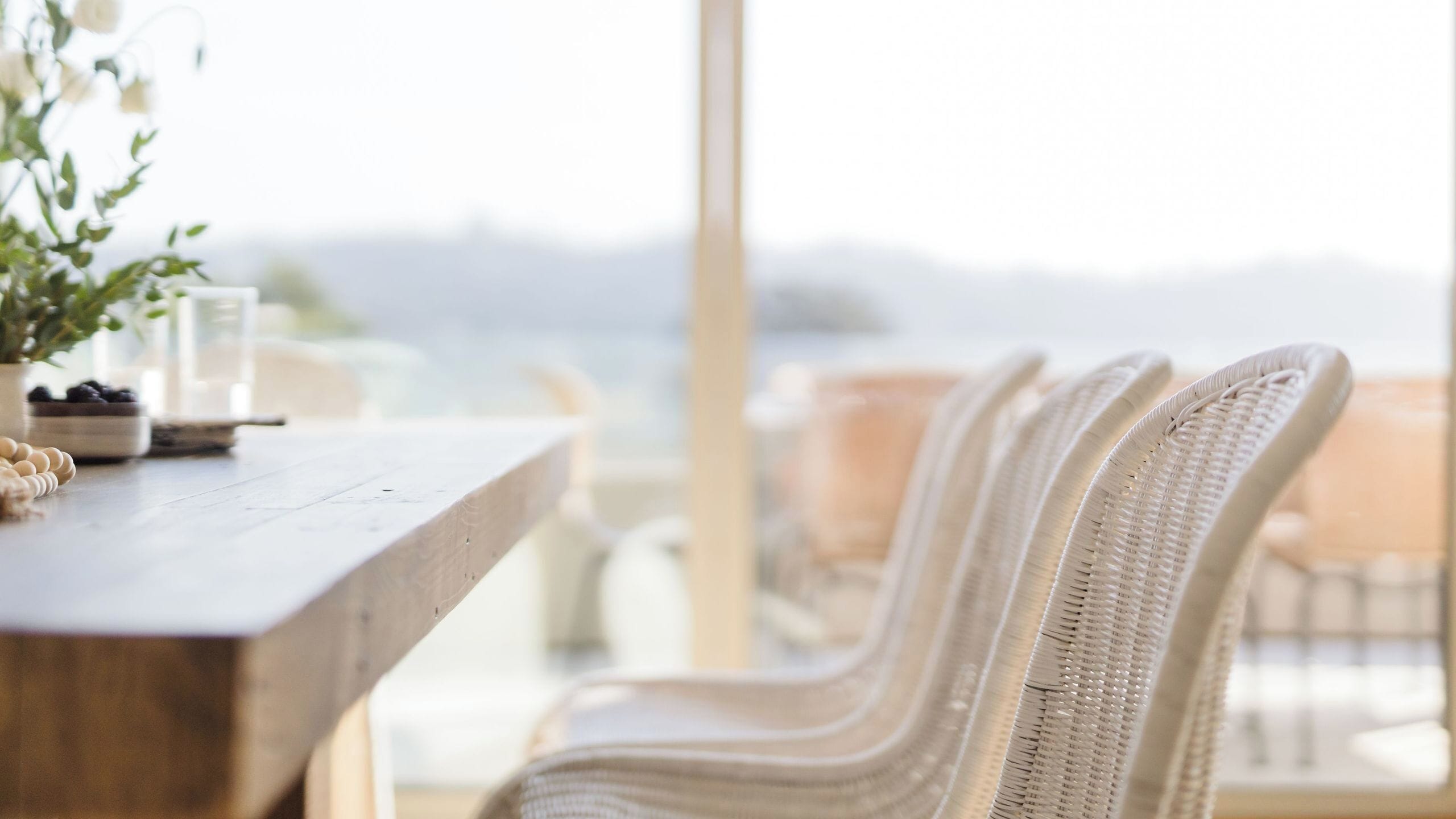 Een houten tafel met een potplant en een schaal fruit. Er staan rieten stoelen bij, met uitzicht op de zee op de achtergrond.