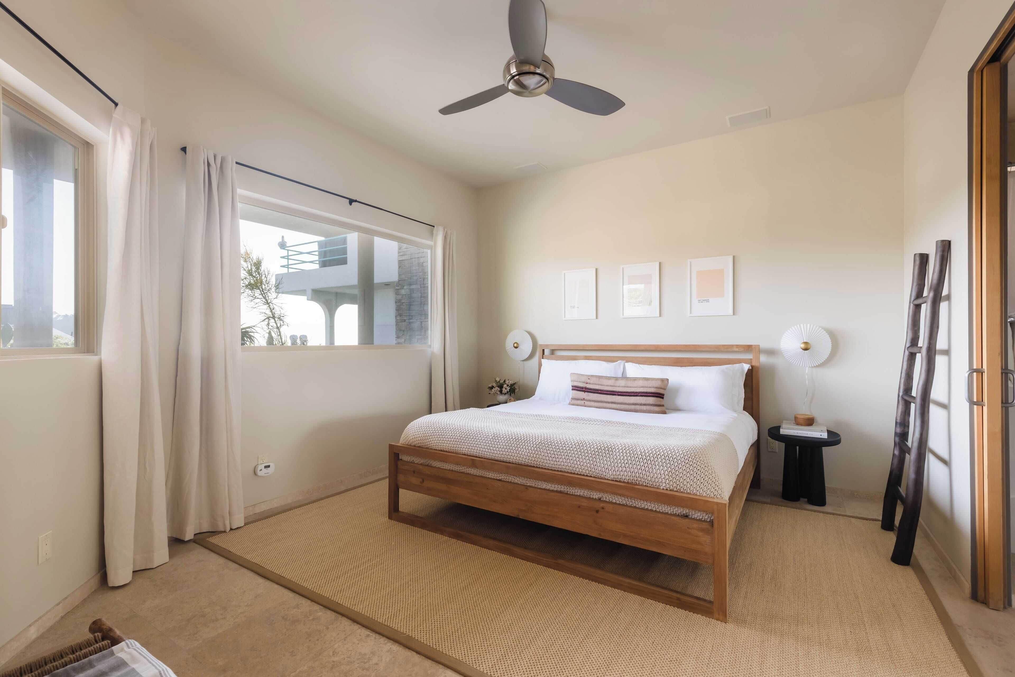 A bedroom with a bed, a ceiling fan, a window with curtains, a small bedside table, and wall-mounted picture frames.