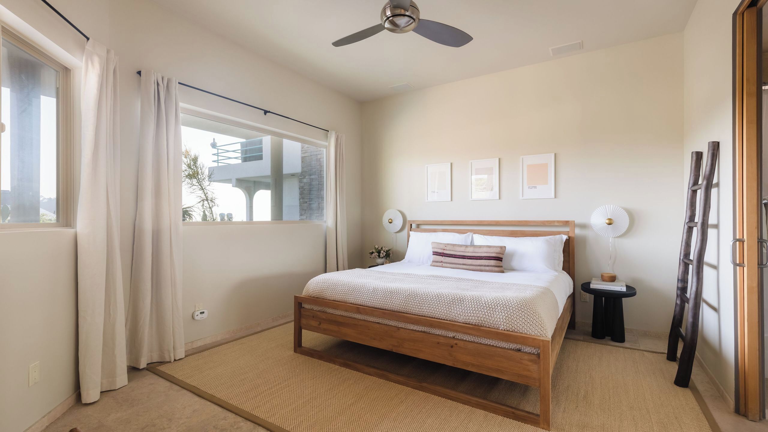 Una camera da letto con un letto, una ventola a soffitto, una finestra con tende, un comodino con una lampada e un quadro appeso al muro.