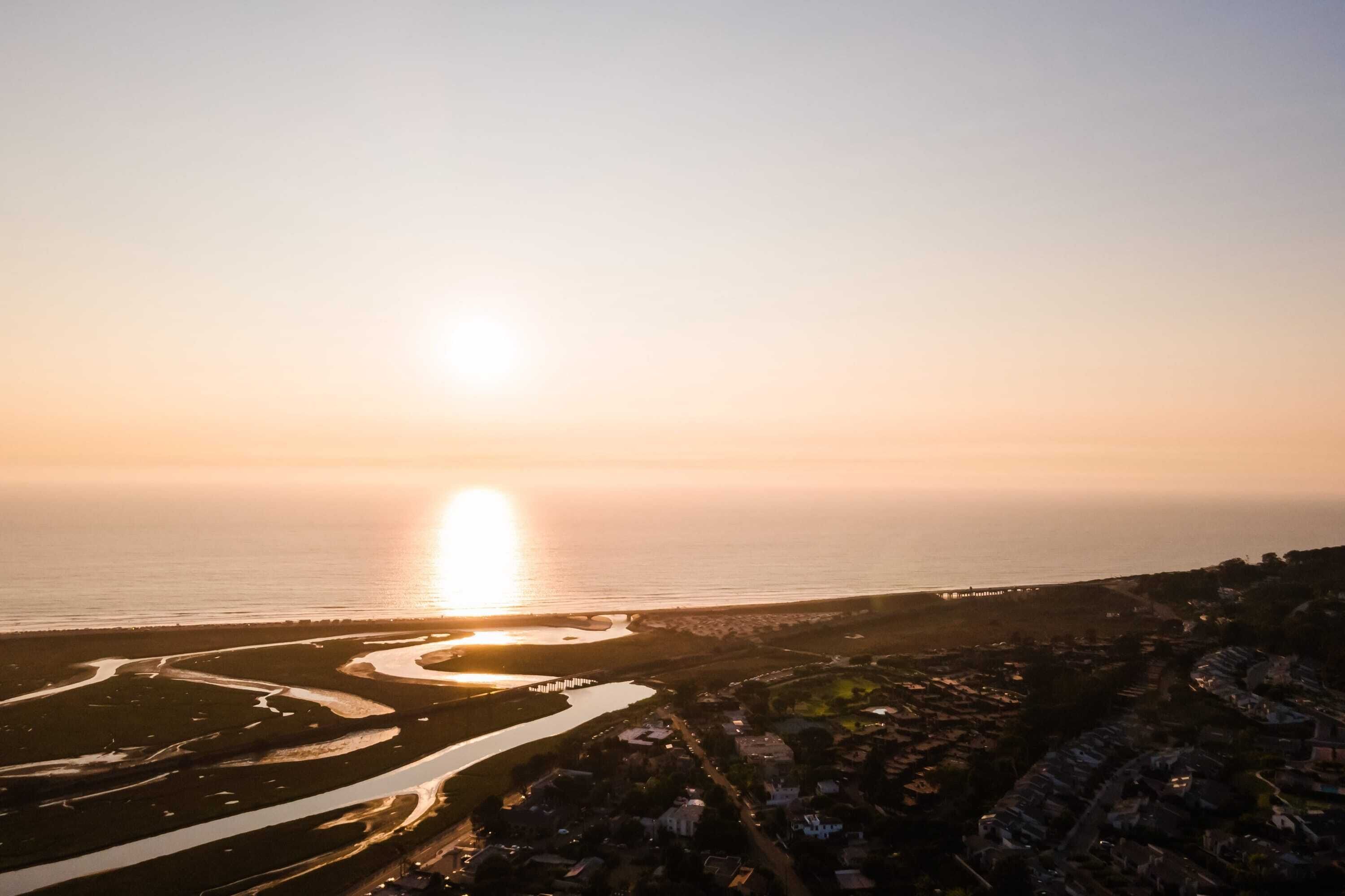 Luchtbeeld van een kustplaats in de zonsondergang, met een rivier, wegen en woonwijken.