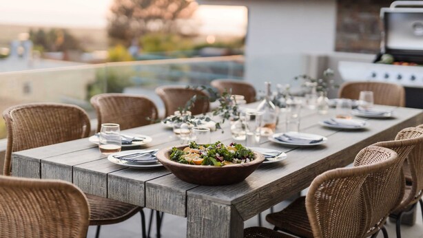 A dining table set outdoors with wicker chairs, a bowl of salad, and a grill in the background. - Seascape by Avantstay Spacious Contemporary Oasis in Del Mar (Del Mar)