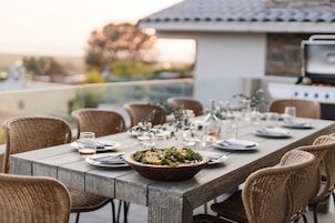 Een eettafel buiten, met rieten stoelen, een saladekom en een barbecue op de achtergrond.