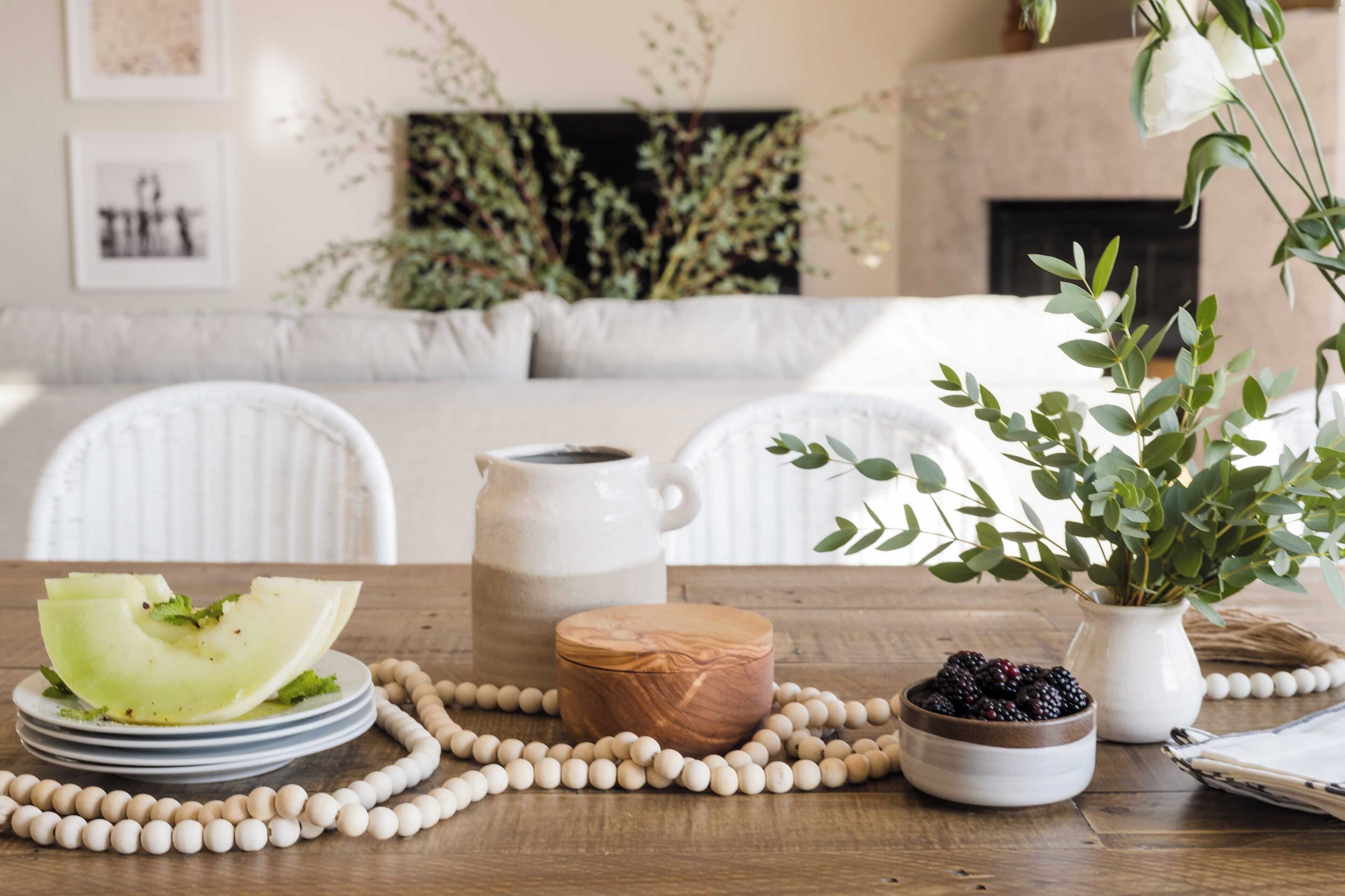 A dining table with a plate of food, a pitcher, a wooden bowl, and a vase with greenery.